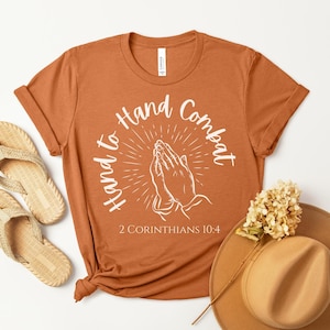 May include: An orange t-shirt with the text "Hand to Hand Combat" and an illustration of praying hands. The shirt also features the Bible verse 2 Corinthians 10:4. The shirt is paired with a pair of woven sandals and a brown hat.