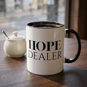 May include: A white ceramic mug with the text "HOPE DEALER" in black. The mug has a black handle and rim, and contains a dark liquid, possibly coffee. A small white sugar bowl with a lid and spoon are next to the mug.