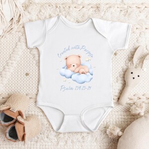 May include: White baby onesie with a watercolor illustration of a teddy bear on a cloud, with the text "Created with Purpose" and "Psalm 139:13-14". The onesie is surrounded by baby accessories, including booties and a stuffed bunny.