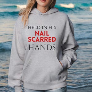 May include: A light gray hooded sweatshirt featuring a front kangaroo pocket and long sleeves. The text HELD IN HIS NAIL SCARRED HANDS is printed on the chest, with the words NAIL SCARRED in red and the remaining words in black lettering.