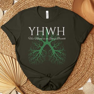 Pode incluir: T-shirt verde escuro com o texto branco "YHWH" e a frase "His Name is in Every Breath". Um gráfico verde em forma de árvore está abaixo. A t-shirt é exibida numa superfície tecida com um chapéu castanho.