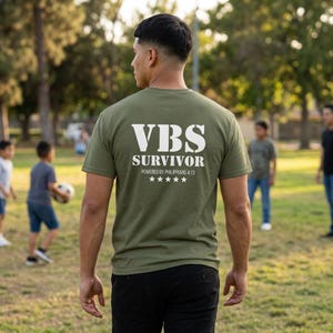 Könnte beinhalten: Ein olivgrünes Kurzarm-T-Shirt mit weißem Aufdruck auf der Rückseite, der VBS SURVIVOR, POWERED BY PHILIPPIANS 4:13 und eine Reihe von fünf weißen Sternen zeigt. Das T-Shirt wird von einer Person getragen, die von hinten zu sehen ist.