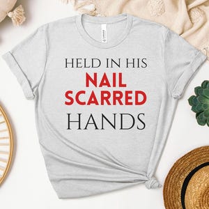May include: A light gray short-sleeve t-shirt featuring the black and red text HELD IN HIS NAIL SCARRED HANDS printed on the front. The shirt has a crew neckline and a standard fit, showcasing a clear graphic design centered on the chest area.