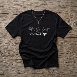 Può includere: Una t-shirt nera a maniche corte con la scritta bianca Father Son Spirit. Sotto il testo ci sono tre icone grafiche bianche: una corona, una corona di spine e una colomba. Una collana color oro con un ciondolo a croce poggia sullo scollo.