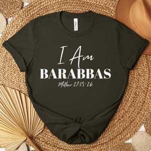 May include: Dark olive green t-shirt with the text "I Am Barabbas" in white script and block letters. The Bible verse Matthew 27:15-26 is printed below. The shirt is styled on a woven mat with a brown hat and decorative elements.