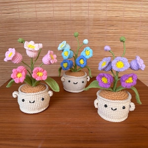 May include: Three crocheted flower arrangements in small pots. Each pot has a smiling face and contains a cluster of colorful flowers. The flowers are pink, blue, and purple with yellow centers and green leaves. The pots are beige.
