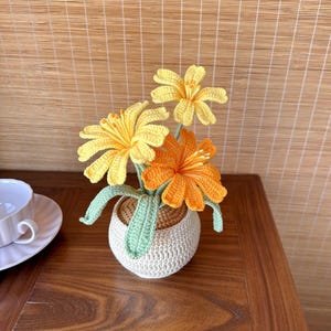 May include: A handmade crochet flower arrangement featuring three vibrant flowers in shades of yellow and orange. The flowers are set in a small, off-white crocheted pot with green leaves. The arrangement is placed on a wooden surface.