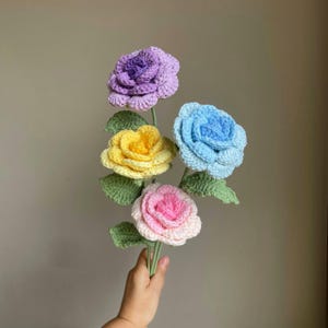 Puede incluir: Un ramo de cuatro rosas de ganchillo en tonos morado, amarillo, azul y rosa. Cada flor tiene hojas y tallos verdes. Las flores hechas a mano se sostienen en una mano.