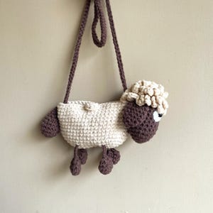 May include: A crochet sheep-shaped bag with a long strap. The bag is mainly off-white with brown details on the head, legs, and strap. The sheep has a fluffy, textured head and a small black eye. The bag is designed to be worn over the shoulder.
