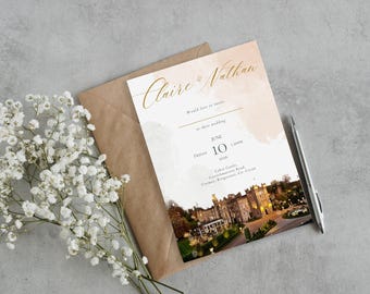 May include: A wedding invitation with gold script that reads "Claire & Nathan". The invitation is on a cream background with a watercolor wash and a photo of a castle. A brown envelope, pen, and white flowers are also in the image.