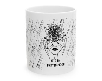 Puede incluir: Taza de cerámica blanca con un dibujo lineal negro de una cara con manos cubriendo los ojos y un garabato enredado encima. El texto "IT'S OK NOT TO BE OK" está debajo. La taza tiene un fondo blanco con marcas de arañazos negros.