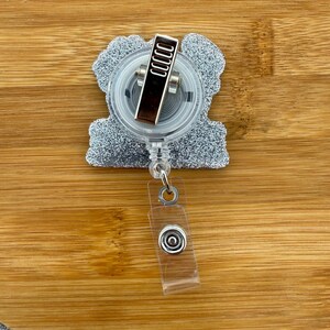 May include: A silver glitter badge reel with a clear plastic retractable cord and a metal clip. The badge reel has a clear plastic snap closure and is set against a wooden background.
