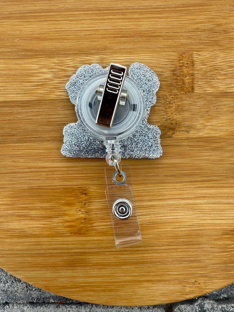 May include: A silver glitter badge reel with a clear plastic retractable cord and a metal clip. The badge reel has a clear plastic snap closure and is set against a wooden background.