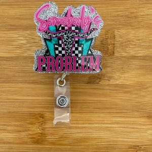 May include: A glittery badge reel with the text "Somebody's Problem" in pink. The design includes a turquoise and black checkered background, a cow skull, and pink lettering. The reel has a clear plastic strap and a metal clip.