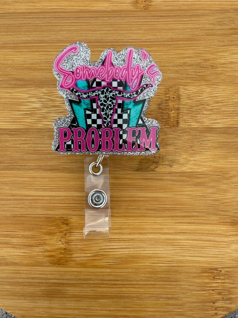 May include: A glittery badge reel with the text "Somebody's Problem" in pink. The design includes a turquoise and black checkered background, a cow skull, and pink lettering. The reel has a clear plastic strap and a metal clip.