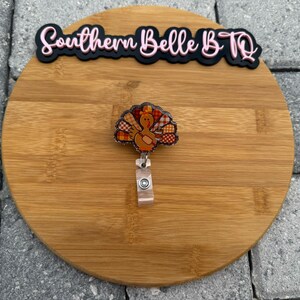 May include: A turkey-themed badge reel with a colorful turkey design. The badge reel is attached to a clear plastic clip. The badge reel is on a round wooden surface. The text "Southern Belle B.TQ" is above the badge reel.