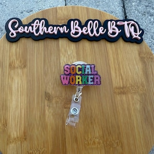May include: A badge reel with the words "SOCIAL WORKER" in rainbow colours with glitter accents. The reel is attached to a clear plastic retractable cord. Above the reel is a black sign with the words "Southern Belle BTQ" in pink.