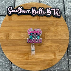 May include: A badge reel with the text "Somebody's Problem" in pink and teal, with a checkered flag design. The reel is attached to a clear plastic holder. The words "Southern Belle B.TQ" are above the reel.
