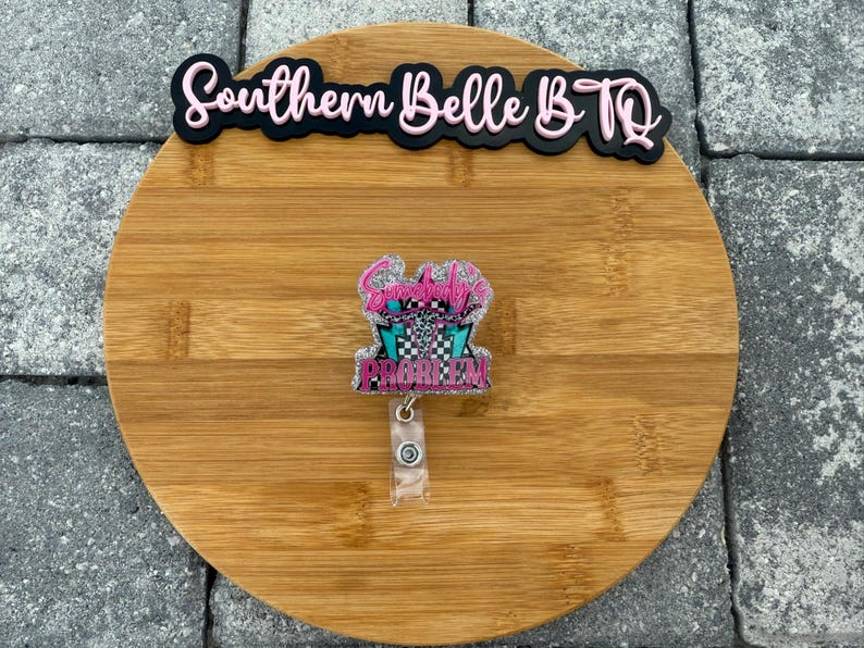 May include: A badge reel with the text "Somebody's Problem" in pink and teal, with a checkered flag design. The reel is attached to a clear plastic holder. The words "Southern Belle B.TQ" are above the reel.