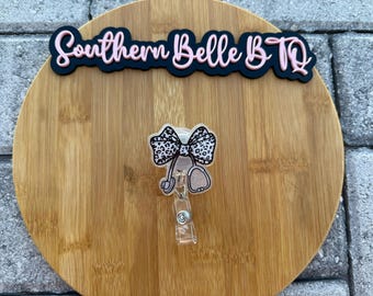 Puede incluir: Un carrete de insignia con un diseño de lazo y estetoscopio. El lazo tiene un estampado de animales en blanco y negro. El carrete tiene un clip de plástico transparente. El texto "Southern Belle BTO" está arriba.