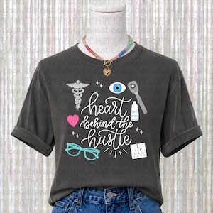 Può includere: Una t-shirt grigio antracite a maniche corte con la scritta bianca "heart behind the hustle" circondata da grafiche a tema medico tra cui un caduceo, un occhio blu, un oftalmoscopio, un cuore rosa, collirio, occhiali e un ottotipo.