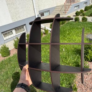 May include: A dark brown, wooden, decorative shelf with a unique, curved design. The shelf has multiple compartments and is held by a person. The shelf is made of wood and has a smooth finish. The shelf is designed to be mounted on a wall.