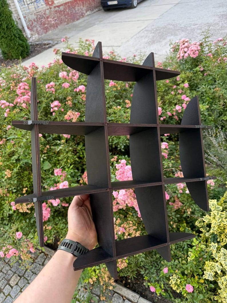 May include: A dark brown, geometric wall shelf with multiple cubby spaces. The shelf is held up by a hand, and is set against a backdrop of pink flowers and green foliage. The shelf is made of wood and has a grid-like design.