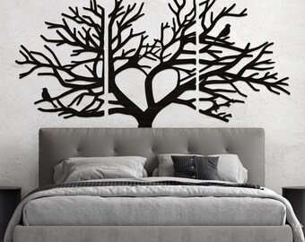 May include: Black metal tree wall art with a heart-shaped design in the center. The art features birds perched on branches and is mounted above a gray bed with pillows and a comforter. The wall is white.