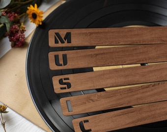 May include: Five horizontal wooden strips with a wood grain texture, each featuring a single black cutout letter. The letters arranged from top to bottom spell out the word MUSIC. The strips are positioned over a black vinyl record.