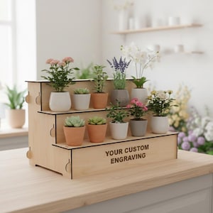 Puede incluir: Un expositor de plantas de madera de tres niveles que muestra varias plantas en macetas, incluyendo suculentas, plantas con flores y hierbas. El expositor tiene un acabado de madera clara y presenta el texto "YOUR CUSTOM ENGRAVING".