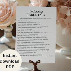 May include: A white card with the text "Wedding Table Talk" and questions, held by a decorative bronze stand. The card is in front of a vase with pink roses and other flowers. A circular "Instant Download PDF" graphic is at the bottom.