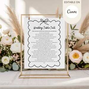 May include: A white paper with black text, titled "Wedding Table Talk," is displayed in a gold metal frame. The text is surrounded by a black, scalloped border. The background features flowers and decorative elements.