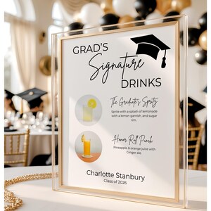 May include: A clear acrylic sign with a gold frame featuring the text Grad's Signature Drinks, a graduation cap icon, and two drink illustrations. The sign lists The Graduate's Spritz and Honor Roll Punch, with the name Charlotte Stanbury and Class of 2026.