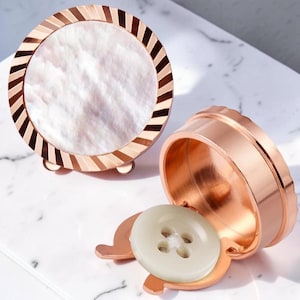 May include: A decorative round box with a mother-of-pearl inlay and a copper-colored metal frame. A matching copper-colored container holds a large, off-white button. The box is resting on a white surface.