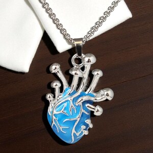 Puede incluir: Un collar con colgante plateado con un dise&ntilde;o en forma de coraz&oacute;n azul y detalles de hongos plateados. El colgante cuelga de una cadena plateada. El collar se muestra sobre una superficie marr&oacute;n oscuro.