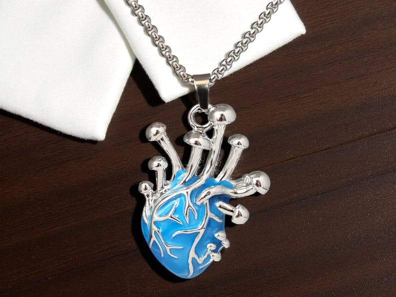 Puede incluir: Un collar con colgante plateado con un dise&ntilde;o en forma de coraz&oacute;n azul y detalles de hongos plateados. El colgante cuelga de una cadena plateada. El collar se muestra sobre una superficie marr&oacute;n oscuro.