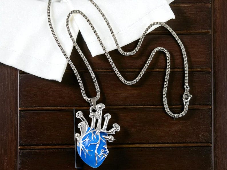 Puede incluir: Un collar plateado con un colgante en forma de coraz&oacute;n anat&oacute;mico azul. El colgante en forma de coraz&oacute;n tiene venas plateadas y una cadena plateada. El collar se exhibe sobre una superficie de madera marr&oacute;n oscuro con una tela blanca.