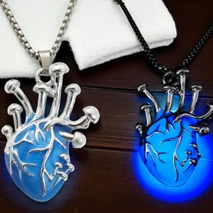 Puede incluir: Dos colgantes en forma de coraz&oacute;n con detalles de hongos. Uno es plateado con un coraz&oacute;n azul, el otro es negro con un coraz&oacute;n azul brillante. Ambos est&aacute;n en cadenas, mostrando un dise&ntilde;o &uacute;nico de inspiraci&oacute;n g&oacute;tica.