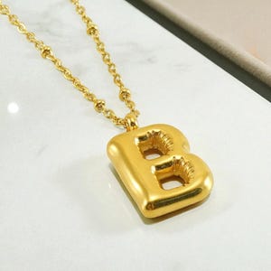 May include: A gold-coloured necklace featuring a pendant shaped like the letter "B". The pendant has a chunky, blocky design. The chain is also gold and has small, round beads. The necklace is shown on a white surface.