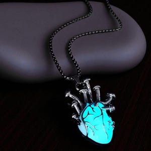 Puede incluir: Un collar plateado con un colgante en forma de coraz&oacute;n humano. El coraz&oacute;n brilla con una luz azul brillante, y el collar tiene una cadena oscura. El colgante tiene un dise&ntilde;o anat&oacute;mico detallado.