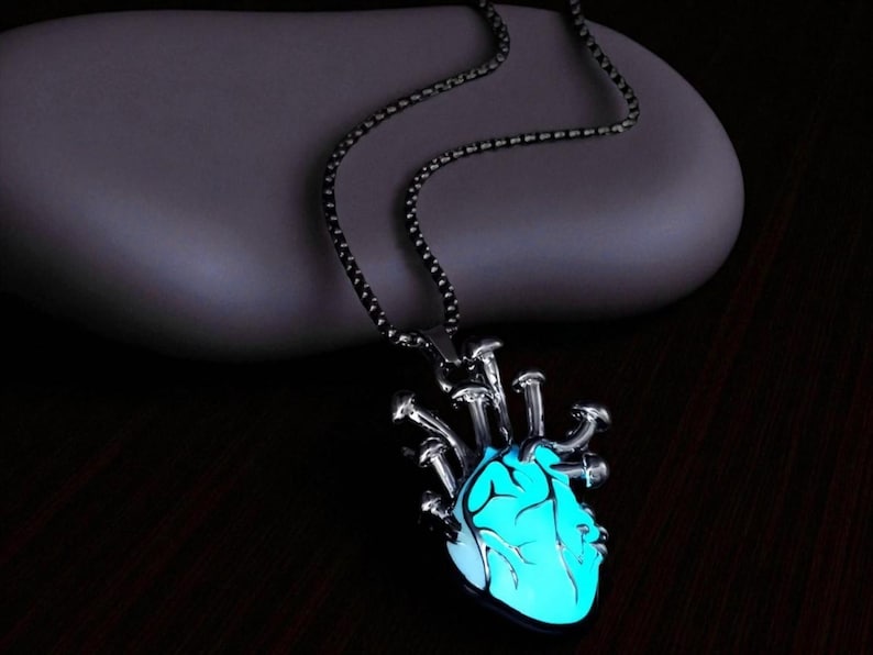 Puede incluir: Un collar plateado con un colgante en forma de coraz&oacute;n humano. El coraz&oacute;n brilla con una luz azul brillante, y el collar tiene una cadena oscura. El colgante tiene un dise&ntilde;o anat&oacute;mico detallado.