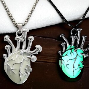 Puede incluir: Dos collares con colgantes en forma de coraz&oacute;n. Uno tiene una cadena plateada y un coraz&oacute;n blanco con detalles de hongos. El otro tiene una cadena negra y un coraz&oacute;n verde brillante con detalles de hongos similares. Ambos est&aacute;n sobre una superficie de madera oscura.