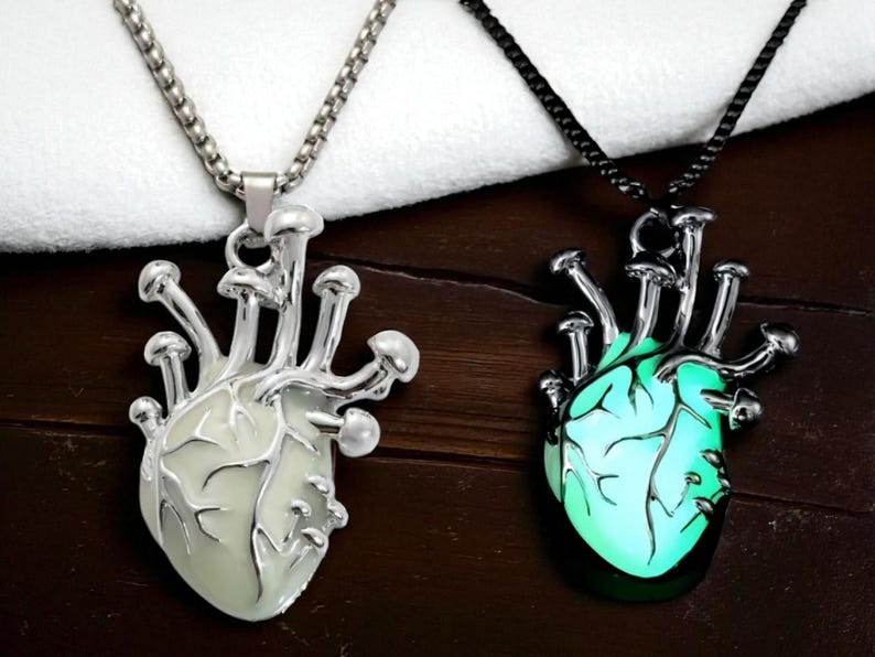 Puede incluir: Dos collares con colgantes en forma de coraz&oacute;n. Uno tiene una cadena plateada y un coraz&oacute;n blanco con detalles de hongos. El otro tiene una cadena negra y un coraz&oacute;n verde brillante con detalles de hongos similares. Ambos est&aacute;n sobre una superficie de madera oscura.