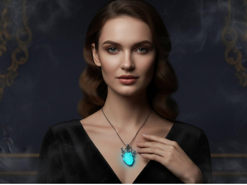 Puede incluir: Una mujer con un vestido negro lleva un collar plateado con un colgante de coraz&oacute;n azul brillante. El colgante es el punto focal, emitiendo un brillo suave y et&eacute;reo contra el fondo oscuro. El collar es una pieza de joyer&iacute;a &uacute;nica.