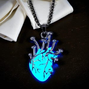 Puede incluir: Collar con colgante plateado con un dise&ntilde;o de coraz&oacute;n azul brillante con detalles en forma de hongo. El colgante cuelga de una cadena oscura, sobre un fondo oscuro con una tela blanca.