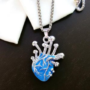 Puede incluir: Collar plateado con un colgante azul en forma de coraz&oacute;n, adornado con detalles plateados en forma de hongo. El colgante cuelga de una fina cadena, sobre un fondo oscuro y tela blanca.