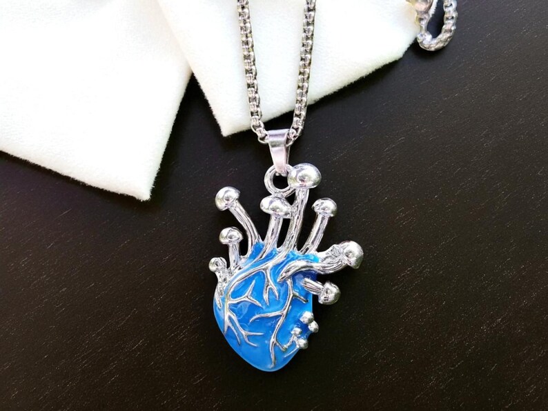 Puede incluir: Collar plateado con un colgante azul en forma de coraz&oacute;n, adornado con detalles plateados en forma de hongo. El colgante cuelga de una fina cadena, sobre un fondo oscuro y tela blanca.