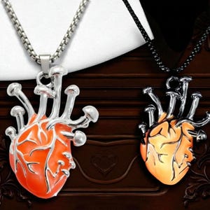 Puede incluir: Dos colgantes en forma de coraz&oacute;n con detalles de hongos. Un colgante es naranja con una cadena plateada, el otro es amarillo con una cadena negra. Los colgantes se muestran sobre un fondo de madera oscura.