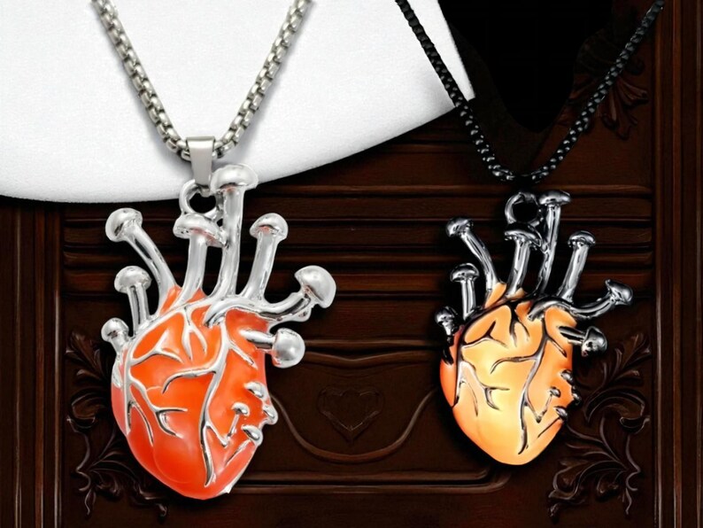 Puede incluir: Dos colgantes en forma de coraz&oacute;n con detalles de hongos. Un colgante es naranja con una cadena plateada, el otro es amarillo con una cadena negra. Los colgantes se muestran sobre un fondo de madera oscura.