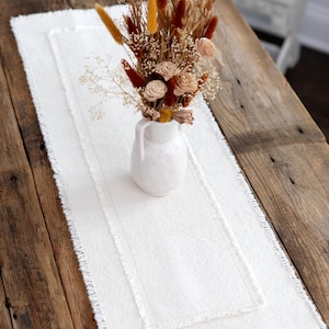 May include: A white linen table runner with a fringed edge, placed on a rustic wooden table. A white ceramic vase holds a dried floral arrangement with brown, beige, and yellow accents. The arrangement adds a touch of natural elegance.
